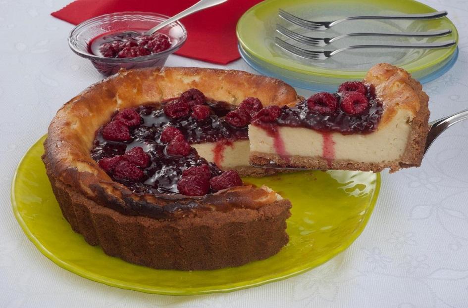 A cheesecake de framboesa é redonda e está sobre um prato redondo de vidro de cor amarela. Um pedaço está sendo retirado, sendo possível ver o interior. Na parte de cima há a geleia de framboesa vermelha e framboesas inteiras.