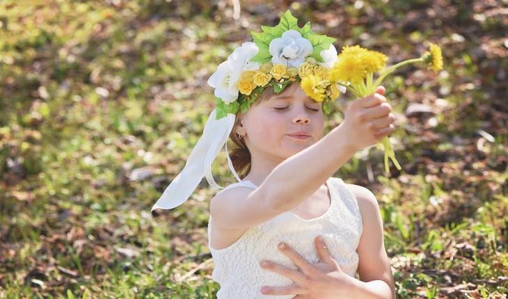 Foto de uma criança de cor branca segurando um pequeno buquê de flores com a mão em seu peito e de olhos fechados. Provérbios de Maio