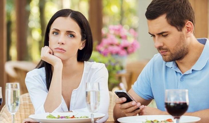 casal em uma mesa, a mulher olha emburrada para a frente enquanto o homem mexe no celular