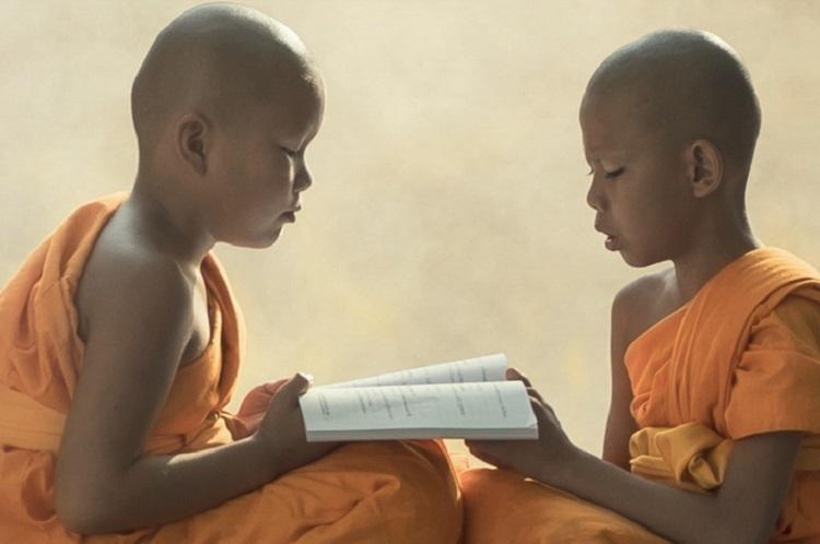 A foto mostra dois meninos sentados com roupas de monge segurando um livro. Eles estão estudando os princípios do Budismo