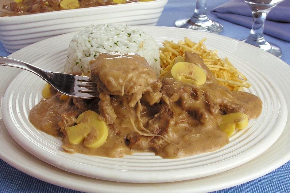 Na foto, o estrogonofe de carne e queijo está em um prato redondo de vidro, acompanhado de arroz branco e batata palha. Um garfo retira parte do estrogonofe.