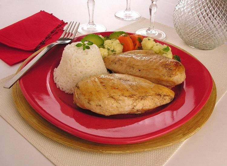 Na foto, o filé de frango com legumes está disposto em prato de vidro vermelho, acompanhado de arroz branco. Os filés estão bem dourados e os legumes estão sortidos, junto à carne. Taças e talheres fazem a decoração ao fundo.