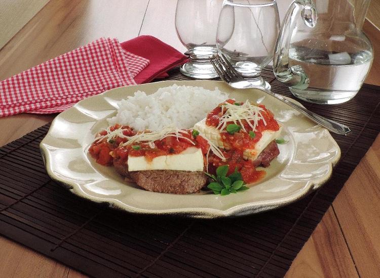 Na foto, o filé-mignon à parmegiana está em um prato de vidro branco. Os filés estão cobertos com molho vermelho e parmesão ralado. A receita está acompanhada de arroz branco.