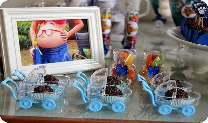 Na foto, há vários carrinhos de bebê miniatura em acrílico na cor azul. Dentro deles há brigadeiros. Junto deles, na mesa, há um porta retrato com a foto da mãe.