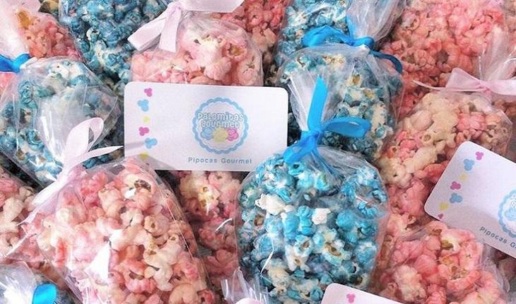 Na foto há vários pacotes de pipoca-doce das cores azul e rosa. Para fechar, há uma etiqueta e fitas da cor das pipocas.