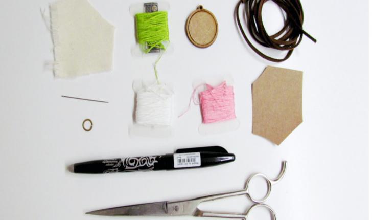 Na foto há materiais de costura para o bordado livre: tecido claro, linhas de costura nas cores verde, rosa e branca, papel cartão, cordão, caneta, tesoura e minibastidores