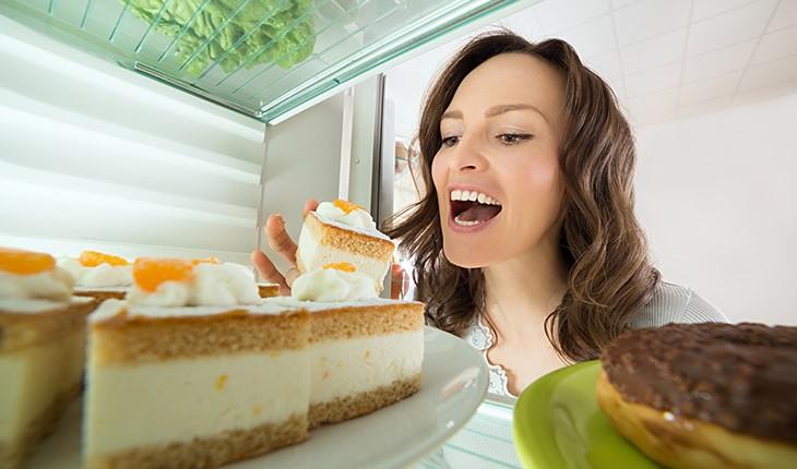 mulher com a geladeira aberta, indo morder um pedaço de bolo