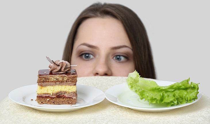A imagem mostra o rosto de uma mulher do nariz para cima. Ela está entre um prato de bolo e outro de salada, mas olhando apenas para o doce