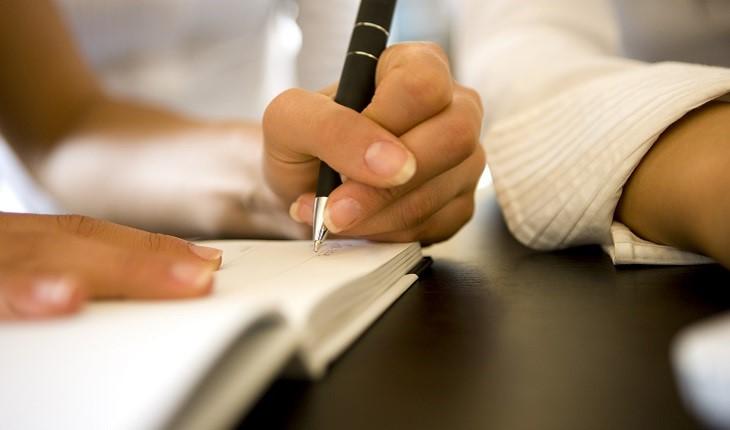 A imagem mostra a mão de uma mulher. Ela está escrevendo em um caderno com uma caneta preta