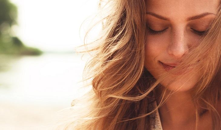 A foto mostra o rosto de uma mulher loira com os olhos fechados e sorrindo. Ela está praticando mindfulness em uma praia