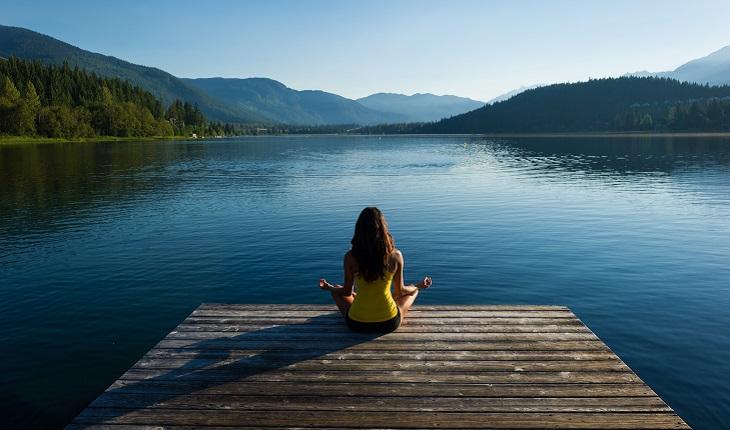Na foto há uma mulher praticando mindfulness sentada na beira de um lago