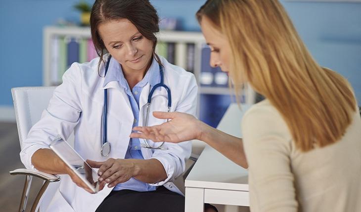 A imagem mostra uma mulher ruiva se consultando com uma médica. A profissional está com um estetoscópio no pescoço e mostrando os exames em um tablet
