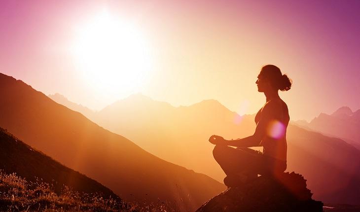 Na imagem, é possível observar uma mulher sentada em um local montanhoso. Ela está praticando mindfulness de manhã