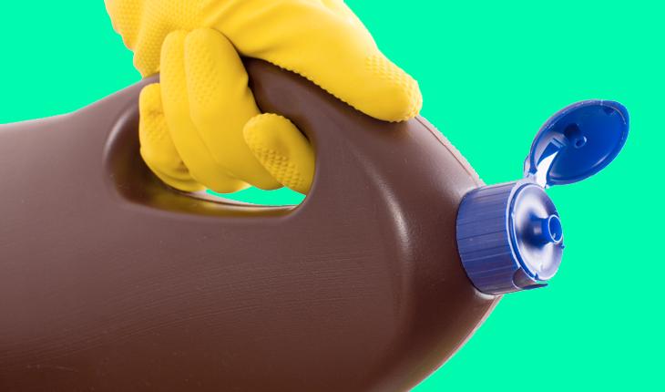 Na foto há um recipiente de água sanitária de cor marrom e é possível ver a mão de uma pessoa com um luva de borracha amarela segurando o recipiente como se estivesse jogando o produto em algum lugar.
