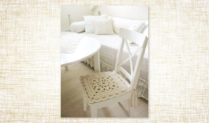 A foto é de uma cadeira branca de madeira que tem uma peça de crochê como assento. A peça é branca e tem cor cru.
