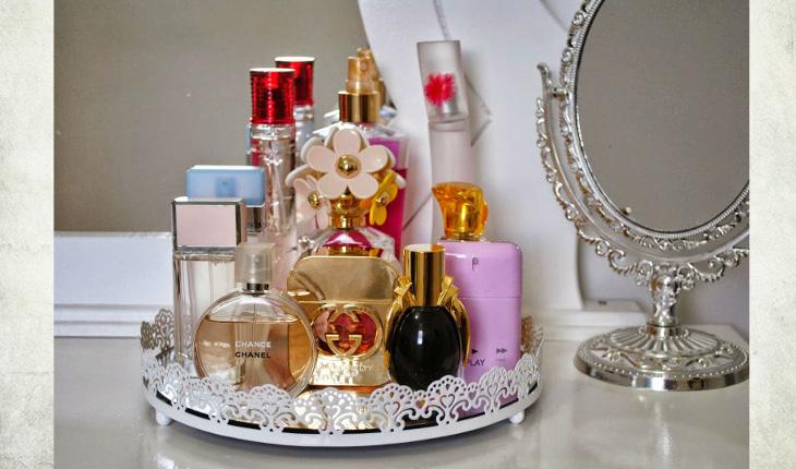 Na foto há uma bandejinha redonda de cor branca em cima de uma bancada e há uma espelho redondo ao lado. Em cima da bandeja há diversos perfumes com recipientes coloridos, como branco, rosa, preto e transparente.