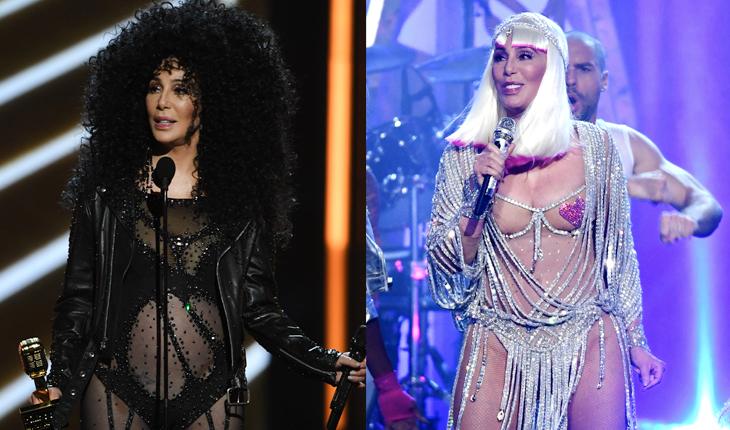 Cher com dois looks bem esquisitos: o primeiro preto e cheio de transparências, com uma peruca de cabelo cacheado bem volumosa. O segundo com franjas prateadas e um coração cobrindo o bico dos seios