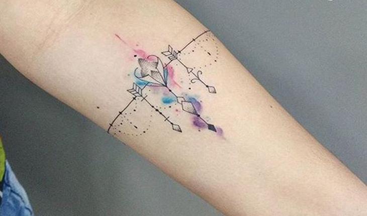 tatuagem de de um bracelete combinado com arabescos, duas flechas e aquerela em rosa, roxo e verde