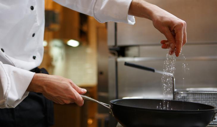 foto das mão de um chef de cozinha segurando uma frigideira e colocando sal na comida