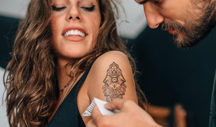 pessoa retirando o papel do decalque da tatuagem feita no braço de uma mulher