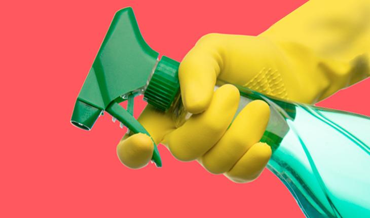 Na foto há um borrifador com tampa verde e vidro verde transparente e é possível ver a mão de uma pessoa com luva de borracha amarela apertando o mecanismo do borrifador.