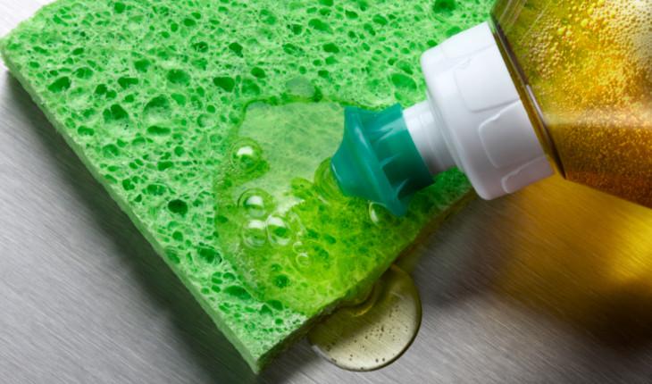 Na foto há uma esponja verde e um recipiente de detergente com tampinha verde está colocando detergente por cima da esponja