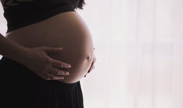 Na foto há uma mulher grávida mostrando a barriga e segurando-as com as mãos.