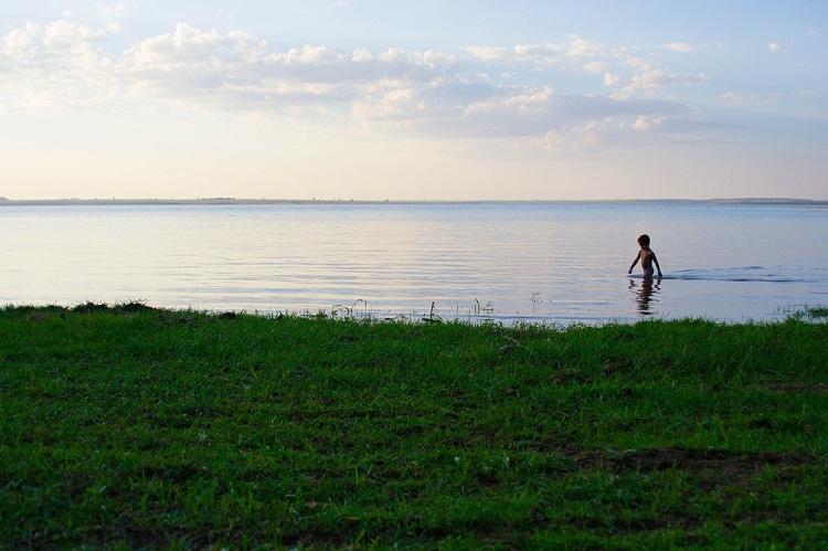Na imagem, um menino indígena está caminhando pelo rio com vegetação verde, lembrando a época de interação com São José de Anchieta.