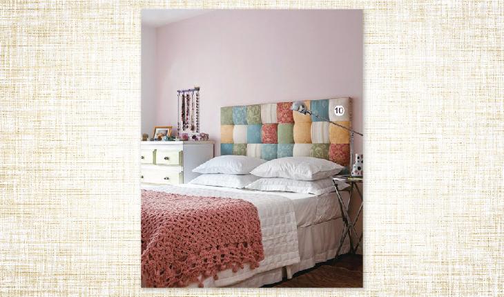 A foto é de uma cama com roupa de cama branca e ao final há uma colha de crochê na cor rosa. A cabeceira da cama é de quadradinhos coloridos com enchimento e é possível ver um criado mudo no lado esquerdo da cama.