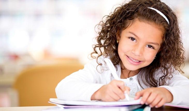 A foto mostra uma menina escrevendo em um papel. Ela está olhando para a câmera e sorrindo