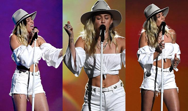 Miley Cyrus usando shortinho banco, com blusinha branca amarrada na frente mostrando a barriga e chapéu