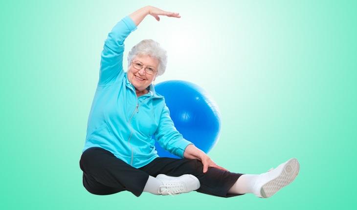 Na imagem, há uma mulher idosa se alongando com uma bola de pilates ao fundo. Os alongamentos são exercícios recomendados na terceira idade