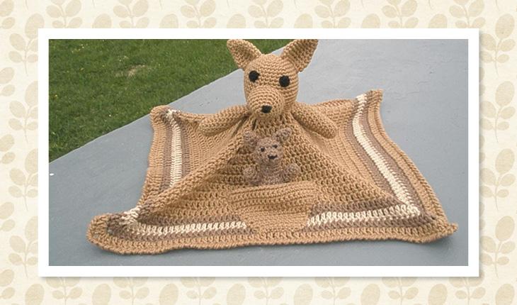 Na foto há uma naninha de crochê de canguru em que, na parte da frente da manta, há um canguru menor. A manta tem cor marrom-claro.