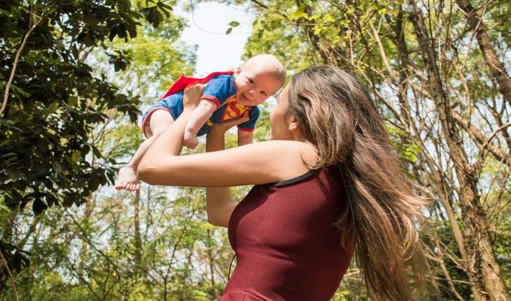 Imagem de uma mãe segurando um bebê em seu colo. A criança está vestida de Super Homem e sorrindo. Proteção para a família