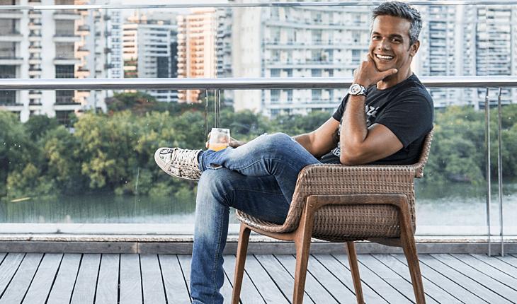 Na lista de famosos que começaram no SBT e fazem sucesso na Globo, está Otaviano Costa. Na foto, o ator e apresentador está sentado em uma cadeira com as pernas cruzadas e, no fundo, uma paisagem urbana