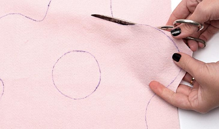 Na foto é possível ver a mão de uma pessoa cortando o feltro rosa ao redor do risco do desenho da cobertura