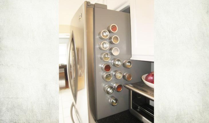 Na foto é possível ver a lateral de uma geladeira em que estão os porta-temperos magnéticos. Há vários porta-temperos e todos estão com algum tipo de tempero, colorido ou não.