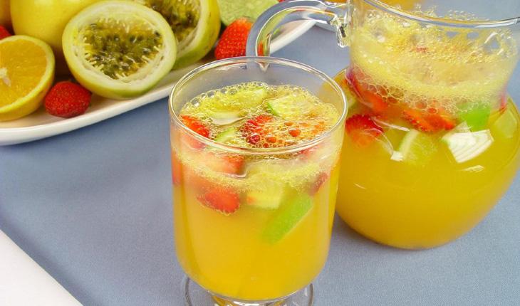 suco com frutas servido em um copo e uma jarra de vidro transparente. O soco é amarelo com pedaços de frutas no topo. Ao fundo, algumas frutas decoram um prato