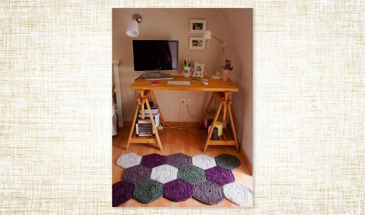 A foto é de um quarto com uma mesinha de madeira com computador e itens de escritório. No chão há um tapete de crochê formado por vários hexágonos das cores cinza, roxo, lilás e branco. O chão é de madeira.
