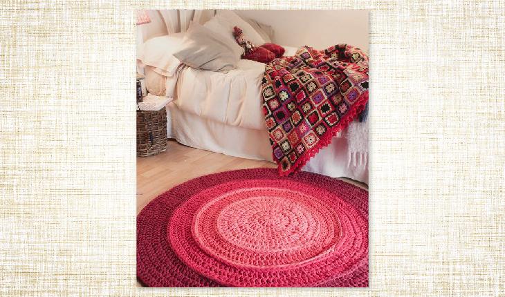 A foto é de um quarto e no chão há um tapete redondo com degradê em rosa de crochê. O chão é claro. Na cama, há uma colcha feita com quadradinhos de crochê e com pequenas flores de crochê em cada um dos quadrados.