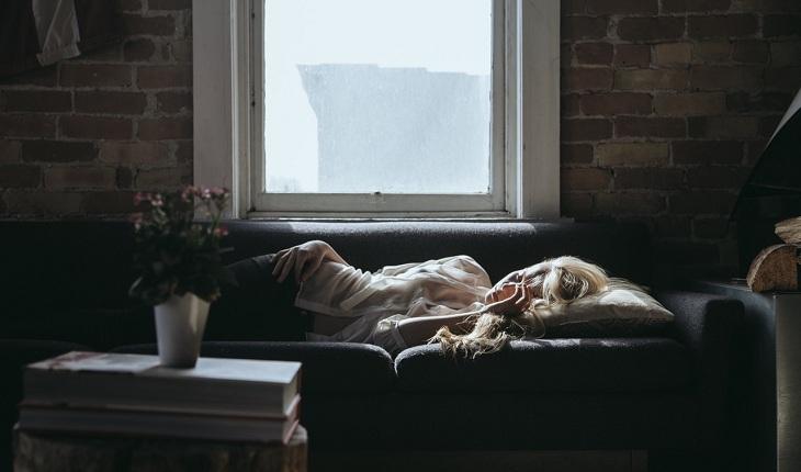 Na imagem, uma mulher está deitada no sofá com cara de dor, dores de tpm, cólica, dor de cabeça. Contraceptivos.