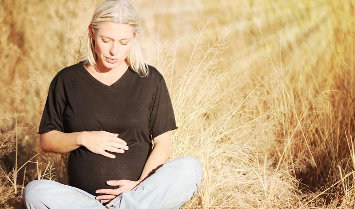 Na imagem, uma mulher grávida está sentada no chão de uma plantação de trigo com blusa preta e calça cinza tocando a barriga. Contraceptivos.