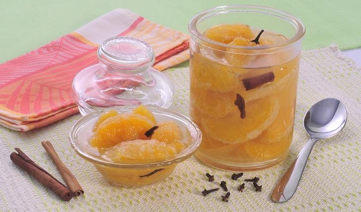 Vidro com compota de laranja com cravos dentro. Ao lado do vidro há um potinho de sobremesa também de vidro com o doce e cravos.