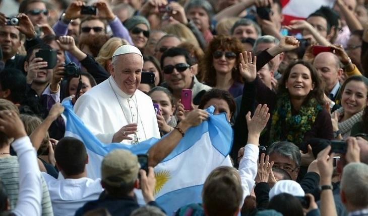 Na imagem, o papa francisco anda pela multidão enquanto alguém acena com a bandeira da argentina esticada. O papa olha com carinho para a bandeira. Dia do papa.