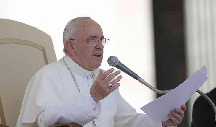 Na imagem, o papa discursa sentado numa cadeira branca segurando os papeis do discurso e com óculos. Dia do papa.