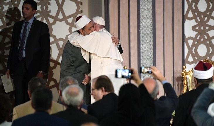 Na imagem, o papa francisco está abraçando e apertando as mãos do líder religioso islâmico em uma conferência com o público tirando fotos deles. Dia do Papa.
