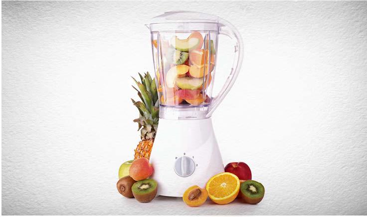 liquidificador com copo transparente, cheio de frutas dentro, frutas ao lado também.