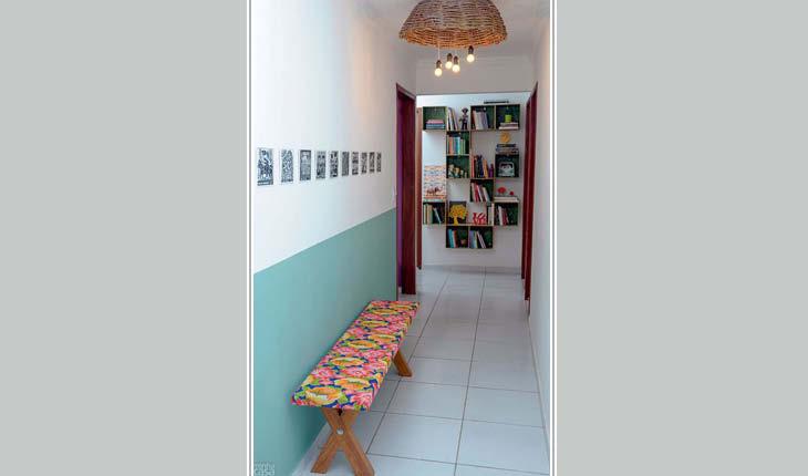 Na foto há um banco de madeira comprido e baixo encostado na parede de um corredor. O tampo do banco é revestido com tecido colorido (chita), deixando-o colorido.