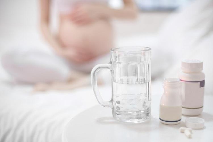 Na imagem, uma mulher grávida está ao fundo fora de foco, no plano da frente, focado, está medicamentos, comprimidos e uma caneca de vidro com água. Para evitar corrimento na gravidez. 