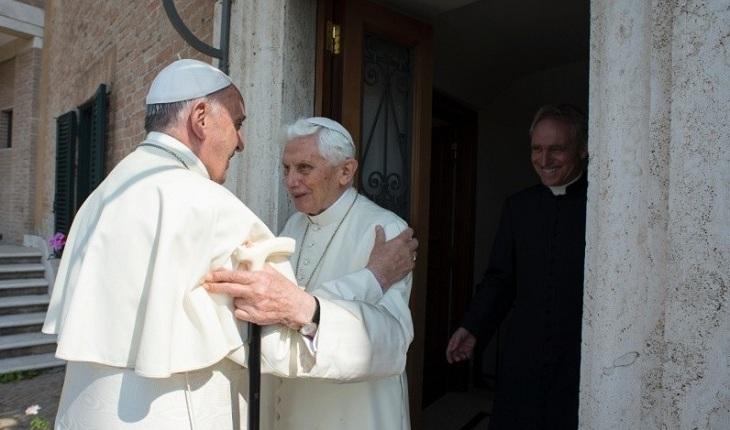 Na imagem, o papa francisco visita o papa bento dezesseis em sua residência e eles se olham e se abraçam com distância. Dia do papa.